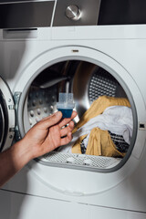 cropped view of man holding measuring cup with blue detergent near washing machine with dirty clothing