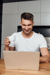 cheerful freelancer holding cup of coffee and looking at laptop