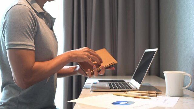 Asian man in casual grey polo shirt using laptop on table and make note on notebook with coffee cup and report chart on table near window with curtain.  Concept for work from home, e learning.