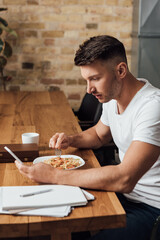 Fototapeta na wymiar Side view of man using smartphone while eating noodles near papers on kitchen table