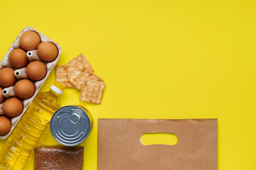 Food supplies bread crumbs, biscuits, buckwheat, eggs, canned goods, sunflower oil paper bag for the period of quarantine on the yellow background. Copyspace. Flatlay