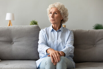 Pensive elderly woman sitting on couch looking away lost deep in her thoughts, she looks worried concerned or lonely, female has senile diseases symptoms memory loss, dementia, mental disorder concept