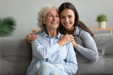 Obraz premium Elderly 65s grandmother sitting on couch holds hand of grown up granddaughter hug her from behind posing looking at camera capture moment, multi-generational relatives family strong connection concept