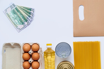 Goods in paper bag, pasta, canned food, eggs, sunflower oil, buckwheat, pearl, grains and us dollar banknotes on white background. Copy space, flat lay. Crisis food stock