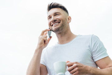 Low angle view of smiling man talking on smartphone and holding cup of coffee isolated on white