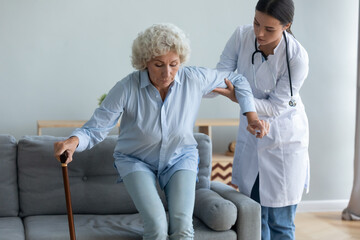 Caring young nurse helping to elderly disabled with walking cane woman get off the couch, age related disease, after illness or operation. Caregiving, provides physical support to old patient concept