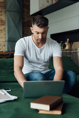 Selective focus of handsome freelancer using laptop near documents and books on couch