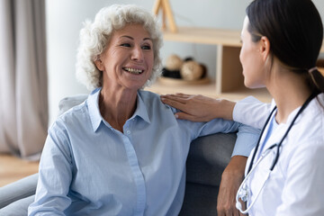 Aged woman talks with young caregiver seated on couch at home, female nurse in white coat puts hand on patient shoulder showing care support and friendly warm relation at visit homecare of old person