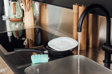 Selective focus of plate, frying pan and sponge in soap near kitchen sink with faucet