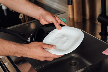cropped view of man holding wet plate and frying pan near sink in kitchen