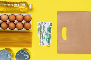 Set of products on yellow background with paper bag and us dollar cash banknotes, flatlay, top view-sunflower oil, chicken eggs, tray, canned, pasta, vermicelli, top view, food, plastic bottle, money