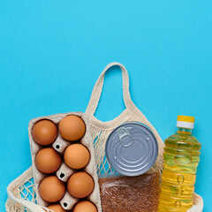 Top view, flatlay. Set of products inside string bag on the blue background. Packaging food from the store. Sunflower oil, eggs, canned food, buckwheat. Delivery and donation concept
