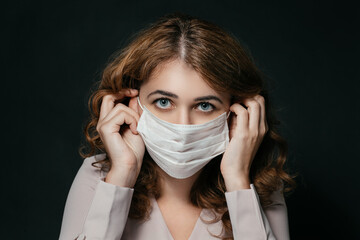 Beautiful young woman wears a respiratory mask, a beautiful female face looks at the camera, a pandemic, a woman's face on a black background, hands in the frame