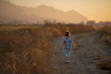 Fototapeta na wymiar Back view on cute little toddler boy in Cyprus Kyrenia, mautains and seaside view walking on the countryside road on sunset. Lifestyle concept