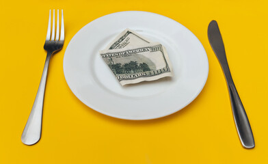 A hundred-dollar bill lies on a white plate between a fork and a knife on a yellow tablecloth. The concept of business lunch, banking, life rentier and financial well-being.