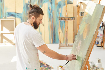 Side view portrait contemporary artist painting picture on easel while working in spacious art studio lit by sunlight, copy space