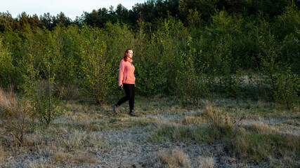 Young woman walking through a meadow carrying a camera, in late evening light.