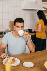 Fototapeta na wymiar selective focus of bi-racial man drinking coffee near woman cooking at home