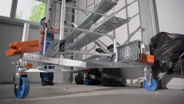 Male Manual Laborer Rolling Metal Scaffolding With Counter Weights To Position It For Job. 
