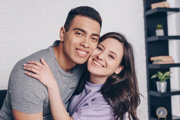 Obraz premium cheerful woman hugging handsome bi-racial man in bedroom