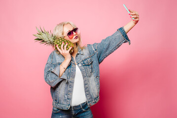 Glamour blond woman in pink sunglasses with pineapple taking selfie on smartphone on color background