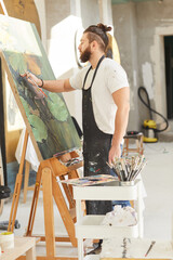 Vertical full length portrait of contemporary bearded artist painting picture on easel while...