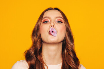young adult girl inflates pink bubble gum burst isolated on yellow background, close up portrait