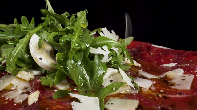closeup moving over macro view beef carpaccio with rocket salad and slices of Parmigiano falling from the sky 4k filmed on black background, Italian food dish