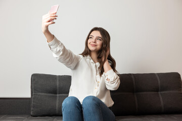 young beautiful brunette woman shooting selfie on her smartphone sitting on the sofa in her...