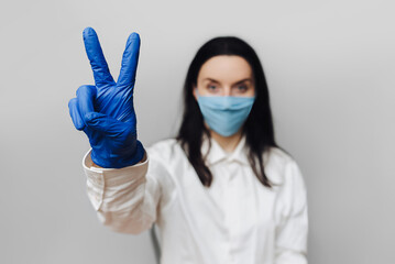 Girl nurse. Young doctor in a white coat, blue protective medical mask and purple rubber gloves shows victory gesture.The concept of the end of quarantine, the pandemic of the coronavirus COVID-19.