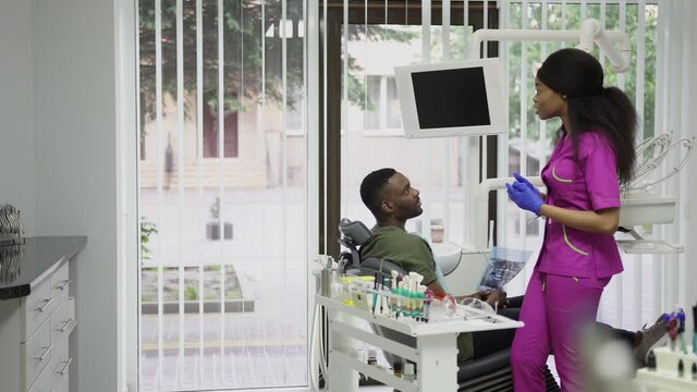 Dentisry concept, healthcare, oral care. Young beautiful african woman dentist discusses the strategy of treatment with the patient, african man, sitting in the chair and holding xray image of jaws