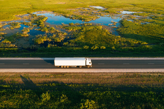 Semi truck with white freight trailer drive on highway in green grass countryside at sunrise, aerial top view. Car with motion blur effect. Road transportation and logistics background