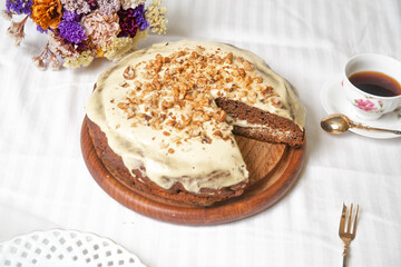 Carrot cake on the white textile material, with flowers, a cup of coffee and gold fork.