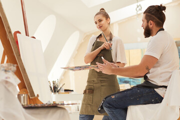 Obraz premium Waist up portrait of smiling young woman holding paintbrush and thinking while standing by blank canvas in art studio lit by sunlight