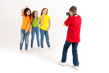Obraz premium A boy photographer takes pictures of three cute girls in bright T-shirts on a white background in the studio. Young photographer and blogger