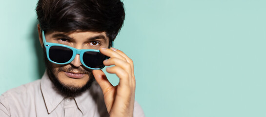 Studio close-up portrait of young guy taking off cyan sunglasses on background of aqua menthe color...