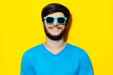 Studio portrait of young guy in blue shirt, wearing cyan sunglasses on background of yellow color.