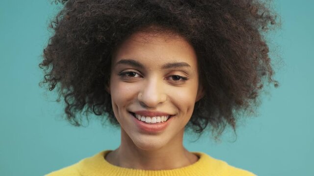 A close-up view of a happy young african american woman with curly hair is opening her eyes laughing and looking to the camera standing isolated over blue wall background in studio