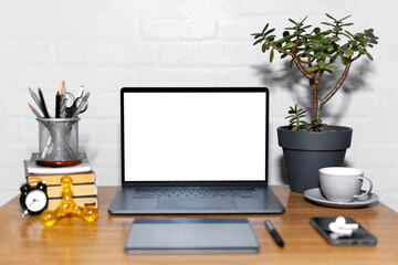 Workplace of freelancer, laptop with graphic tablet on wooden desk beside smartphone, wireless earphones and coffee mug near home flower and books. Background of white brick wall.