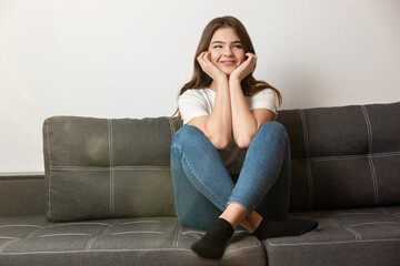 young beautiful smiling brunette woman sitting on the sofa in her appartment holding hands near face looking happy, feels cozy at home