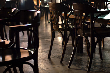 Old retro style empty wooden dining chairs in a dark restaurant with tiled floors and sun rays on the floor