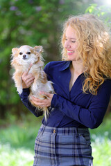 Blond young woman, business dressed, holding a long-haired chihuahua dog outdoor, on a green natural background.