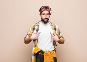 young housekeeper man pointing to self with a confused and quizzical look, shocked and surprised to be chosen against flat wall