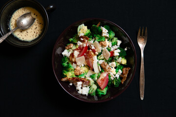 Close-up Broccoli Salad with Bacon, parmesan cheese in a black plate. In the right a silver fork. Top view.