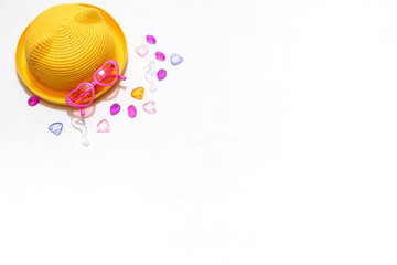 Beach accessories for sun protection: children's yellow straw hat, pink heart-shaped glasses on a white background. Layout with space for text. Ready for travel and beach holidays at sea