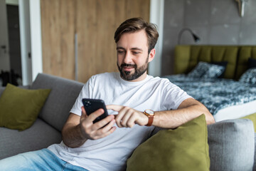 Millennial man sitting in living room on couch at home smiling chatting typing message using cellular. Businessman at home solve business matters by phone. New generation addicted with devices concept