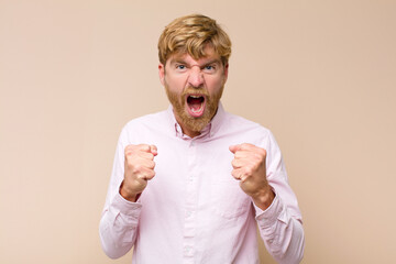 young blonde man shouting aggressively with annoyed, frustrated, angry look and tight fists,...