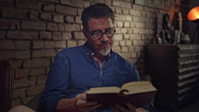 Older man in glasses reading book at home sitting in living room, smiling. 4K video footage.