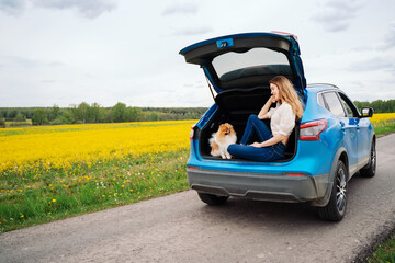 Happy young woman traveler with dog sits in car on spring road. Freedom and traveling couple....