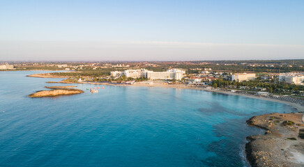 Naklejka na ściany i meble Aerial bird's eye view of famous Nissi beach coastline, Ayia Napa, Famagusta, Cyprus.Landmark tourist attraction islet bay at sunrise with golden sand, sunbeds, sea restaurants in Agia Napa from above